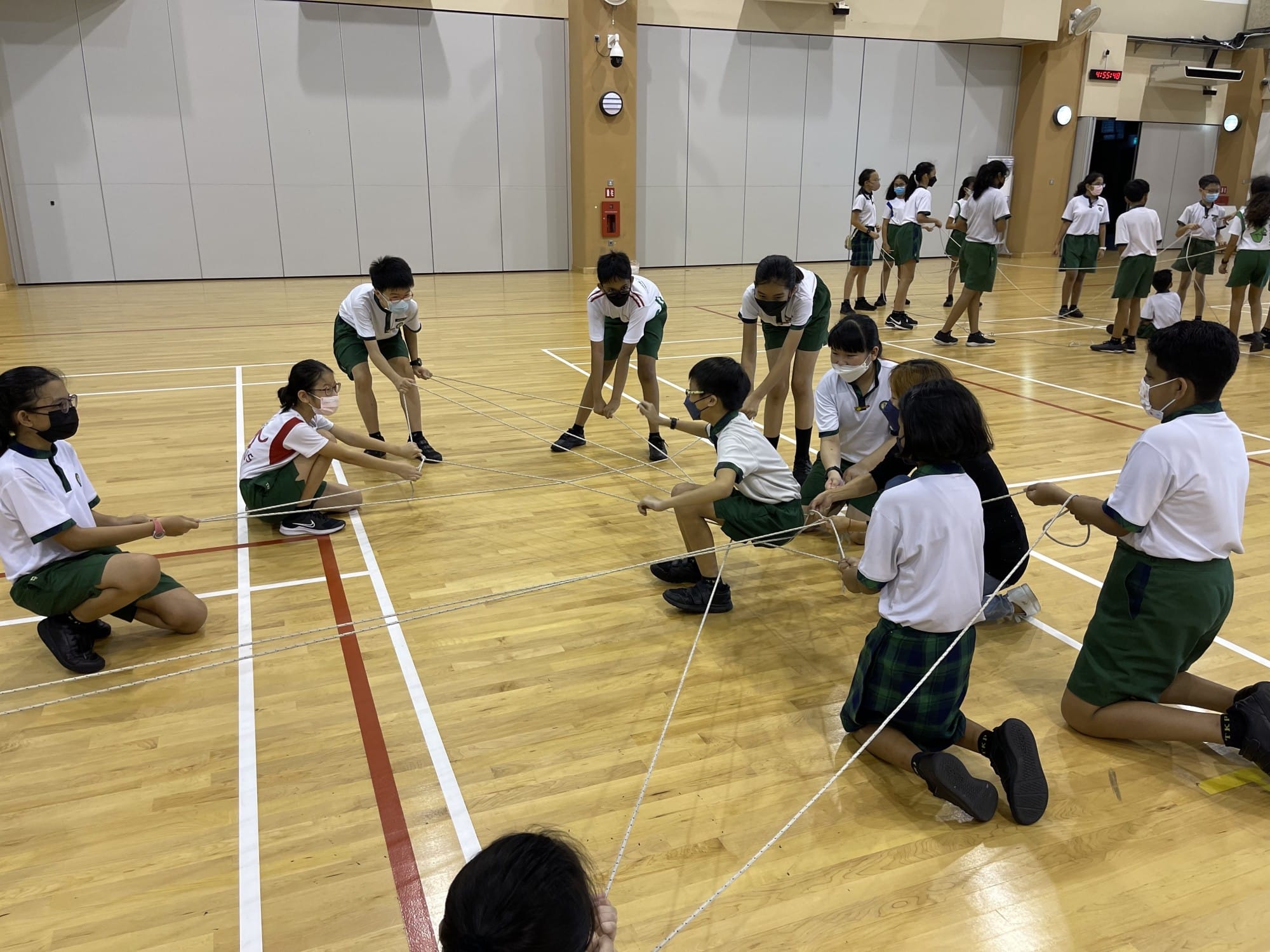Students in green and white uniforms and masks do a rope teamwork activity on an indoor wooden gym floor.